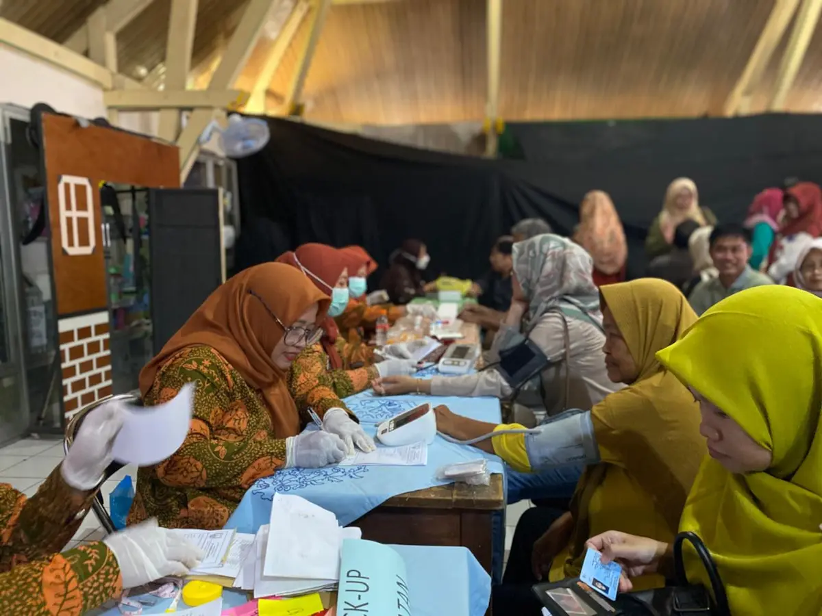 Warga sedang duduk mengantre untuk mengikuti layanan pemeriksaan kesehatan gratis di area halaman sekolah.
